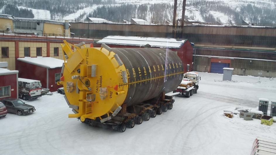 Large steel cylindrical structure mounted on a multi-axle heavy transport trailer outside an industrial workshop in snowy conditions, prepared for offshore or industrial delivery.